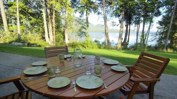Une table ronde en bois, dressée pour quatre personnes avec des assiettes, des couverts et des verres, entourée de chaises, est placée sur une terrasse offrant une vue sur un lac.