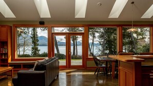 Une pièce spacieuse dotée d’une grande porte-fenêtre coulissante qui donne sur un paysage magnifique, avec un lac et des montagnes en toile de fond. L’intérieur comprend une table à manger en bois avec des chaises, un canapé en cuir noir et un banc en bois.