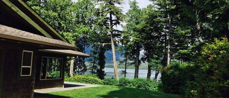 A lodge with a covered porch, a concrete driveway, and a view of a lake surrounded by dense forest.