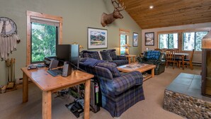 Ein gemütliches Blockhausschlafzimmer mit Holzbalkendecke, einem aufgehängten Hirschgeweih, einem Schreibtisch mit Computern, einer Couch und einem Essbereich.