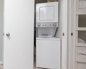 A compact, white laundry room with a washer and dryer stacked vertically, a door with a round handle, and a small shelf with folded towels.