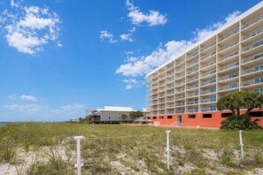 Un hotel frente a la playa con varios balcones, una playa arenosa en primer plano y un cielo azul despejado.