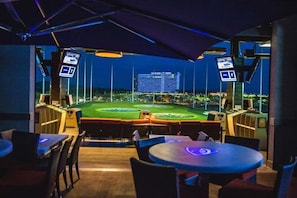 Ein Innenbereich für Sportübertragungen mit Tischen und Stühlen, einem großen Bildschirm, der ein Sportereignis zeigt, und Blick auf ein Stadion im Freien.
