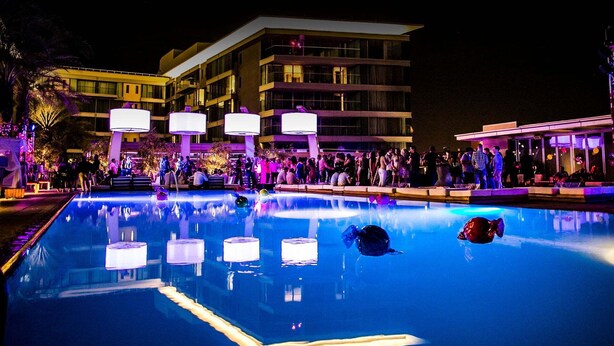 Une piscine entourée de personnes, un bâtiment doté de balcons, et une scène nocturne.