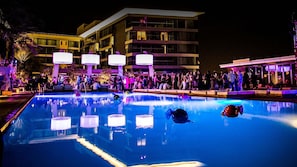 Piscina cercada por pessoas, um edifício com varandas e ambiente noturno.