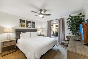 Quarto de hotel com cama, ventoinha no teto, escrivaninha com televisão, planta e janela com cortinas.