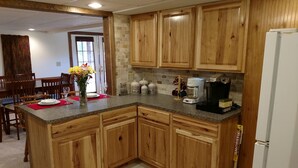 Una cocina con armarios de madera, encimeras de granito y un refrigerador. Se observa un comedor con sillas y una mesa al fondo.