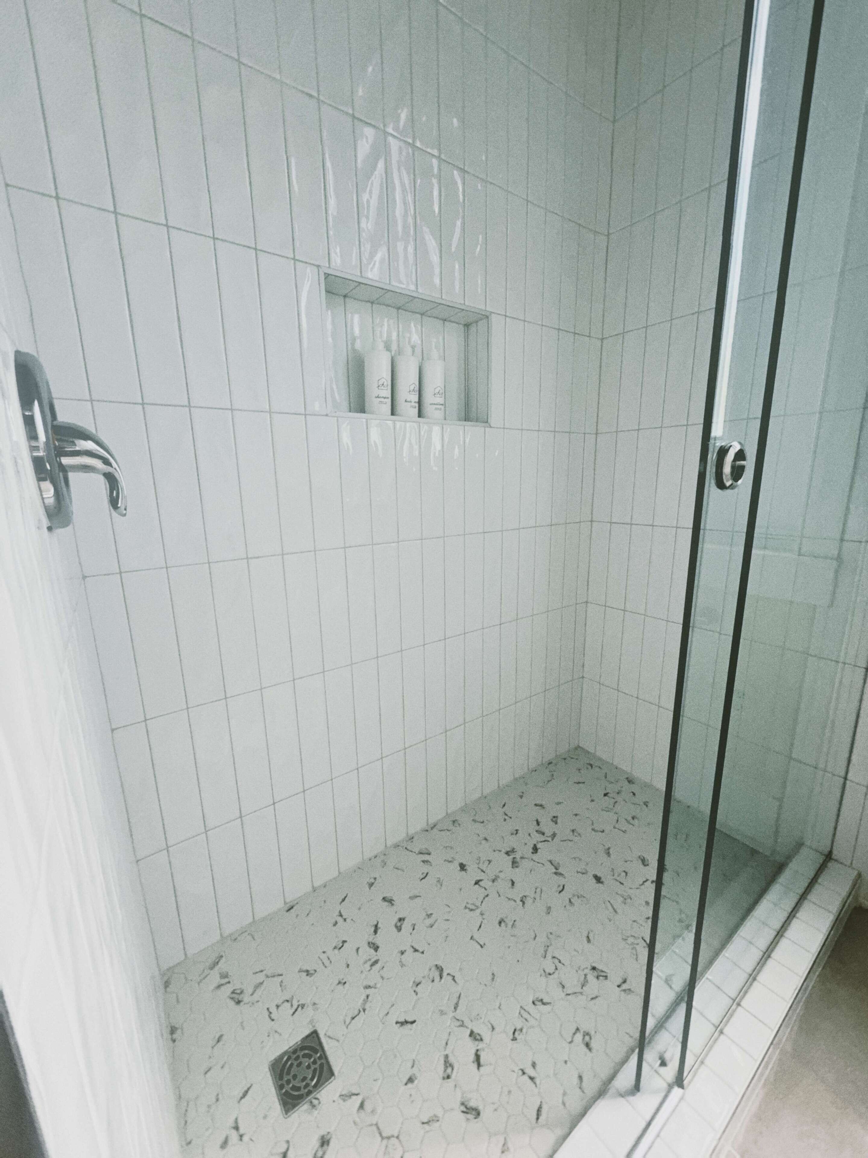 Een douche met een glazen deur, betegelde wanden en een plank met toiletartikelen.