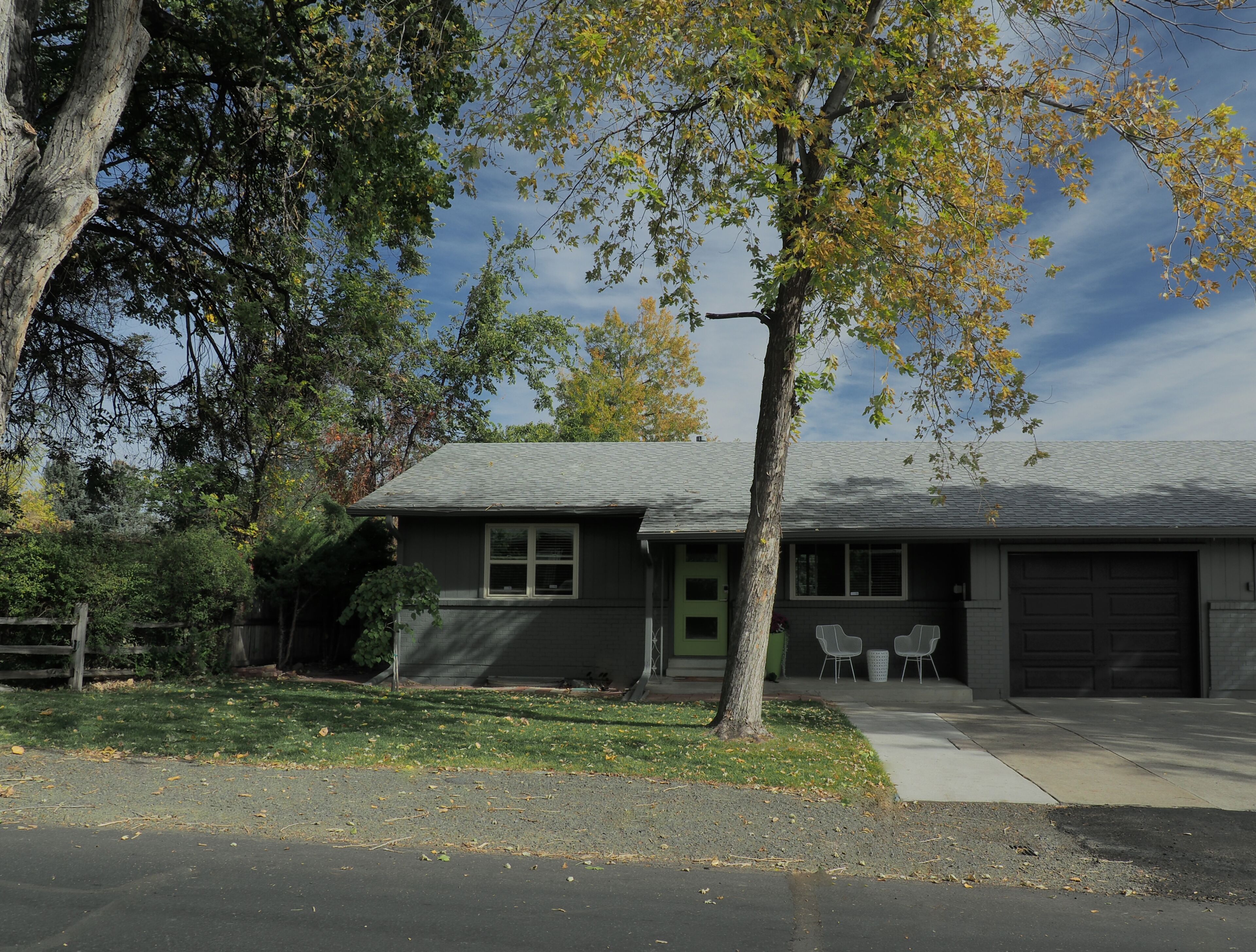 A single-story house with a front garage, a small porch with two chairs, and a tree in front.