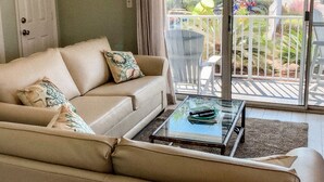 En stue med en beige sofa, et glasbord og udsigt til en balkon med udendørs møbler og grønt.
