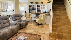 A spacious living room with a brown leather sofa, a beige armchair, and a coffee table with magazines. There is a staircase leading to an upper level.