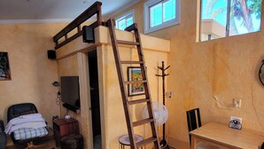 Een kleine kamer met een houten vloer, een houten tafel met twee stoelen, een zwarte bureaustoel, een klein krukje, een houten ladder, een kapstok aan de muur, en een deur naar een andere kamer.