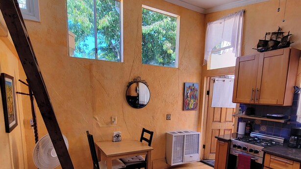 Een keuken met houten kastjes, een eettafel met stoelen, een vloerventilator en een raam met uitzicht op groen.