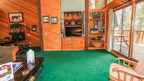 A cozy cabin interior with wooden walls, a green carpet, a fireplace, and a large window overlooking a deck. - Summit Adventure - Walk to Snow Summit, Game Room, Pool Table, BBQ (Big Bear Lake)