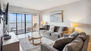 A living room with a glass coffee table, grey sofas, a TV, and a balcony with a view of the sea. - Villa Madeira 206 Beautiful updates|Beach front|Walk to all the activities! (Madeira Beach)