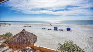 A beach view with a thatched umbrella, lounge chairs, and blue beach chairs. - Villa Madeira 206 Beautiful updates|Beach front|Walk to all the activities! (Madeira Beach)