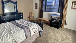 Un dormitorio con cama, cómoda, silla, televisión, ventana y cortinas.