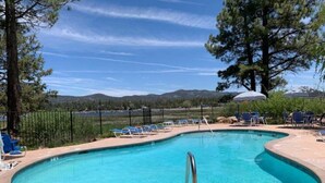 Una piscina azul cristalina con sillones de descanso y sombrillas, rodeada de una valla y árboles, con montañas al fondo.