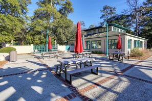 Ein Terrassenbereich im Freien mit Picknicktischen und roten Sonnenschirmen, umgeben von Bäumen und einem Gebäude mit großen Fenstern.