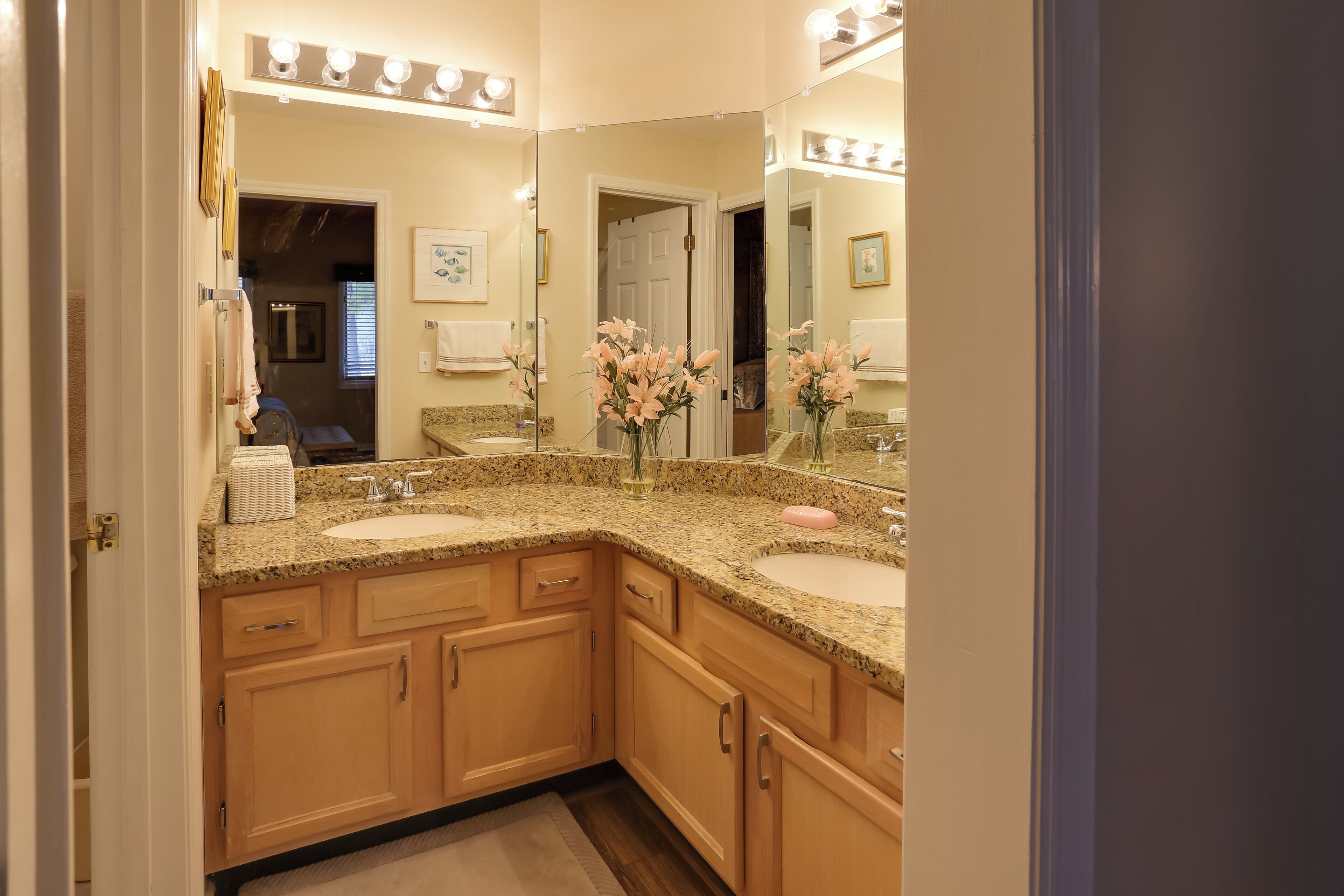A bathroom with two sinks, a vanity with drawers, a mirror with lights, and a vase with flowers.