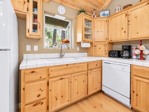 Una cocina con armarios de madera, encimera blanca, un lavabo, un reloj y una ventana con vistas a árboles.