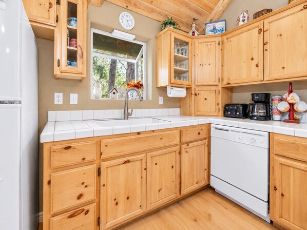 A kitchen with wooden cabinets, a white countertop, a sink, a clock, and a window with a view of trees.