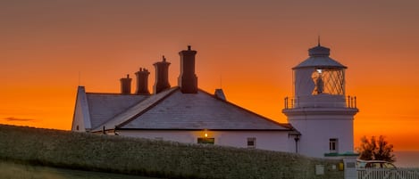 Un phare à l’extérieur blanc, doté d’une lanterne, situé à côté d’un bâtiment comportant de nombreuses cheminées, sous un ciel au coucher du soleil.