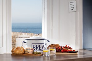 A breakfast setting with bread, a pie, olives, and grapes on a table by a window overlooking the sea.