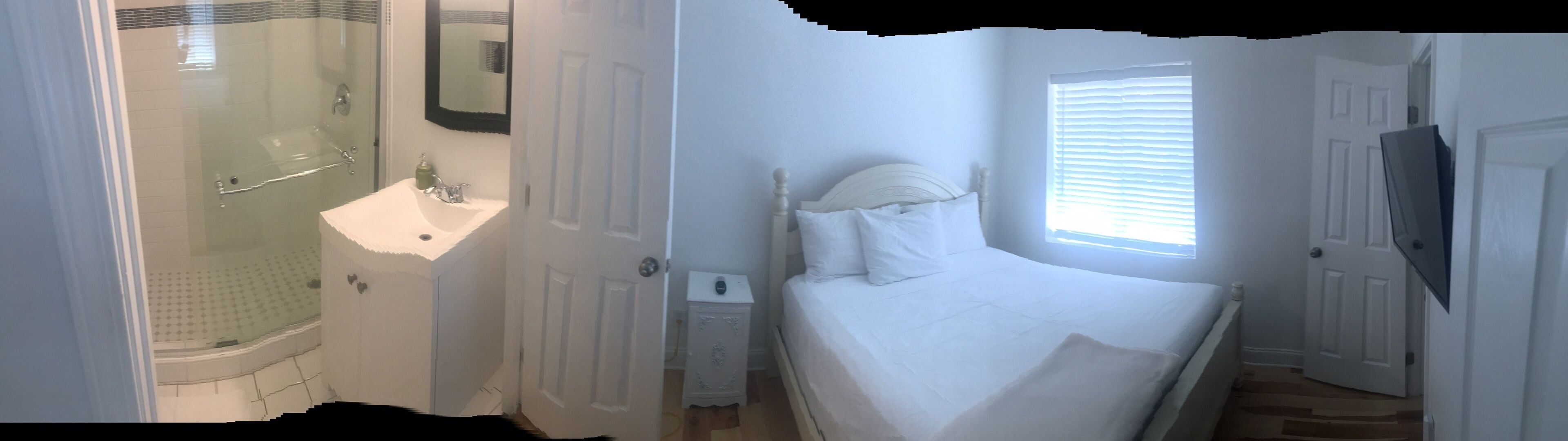 Ein Schlafzimmer mit Bett, Nachttisch, Fenster mit Jalousien, Tür und eigenem Badezimmer mit Dusche und Waschbecken.