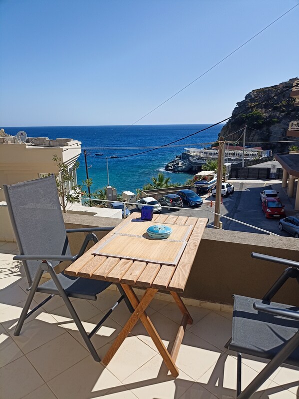 A wooden table with chairs on a balcony overlooking a coastal area with buildings and parked cars.