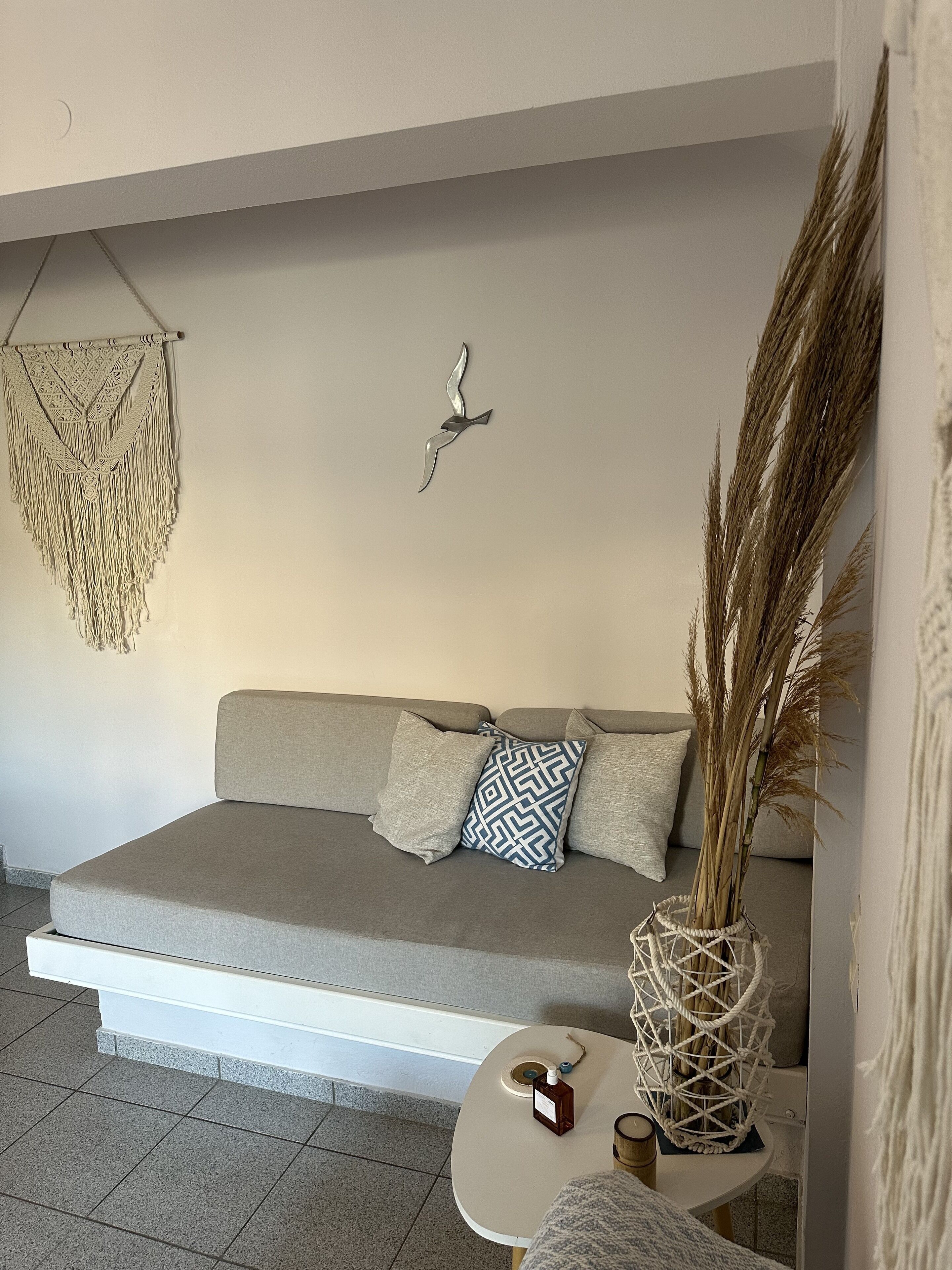Un salon moderne avec un canapé gris, une décoration murale en macramé et un vase contenant des plantes séchées, posé sur une petite table.