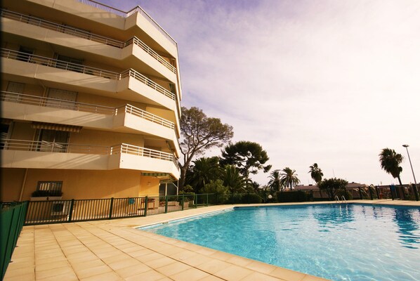 A swimming pool with a tiled deck, surrounded by a fence, and adjacent to a multi-story building with balconies.