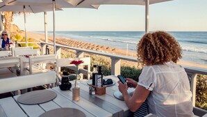 En café ved stranden med udendørs siddepladser, et bord med et kamera, en smartphone og en flaske olivenolie.