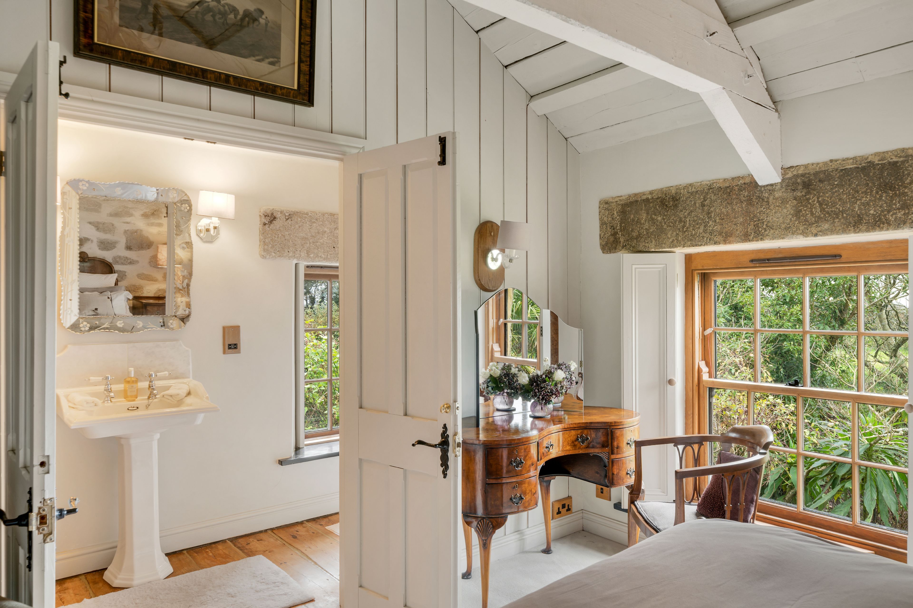 Una habitación con un tocador de madera, un espejo, un lavabo y una ventana con vistas a la vegetación.
