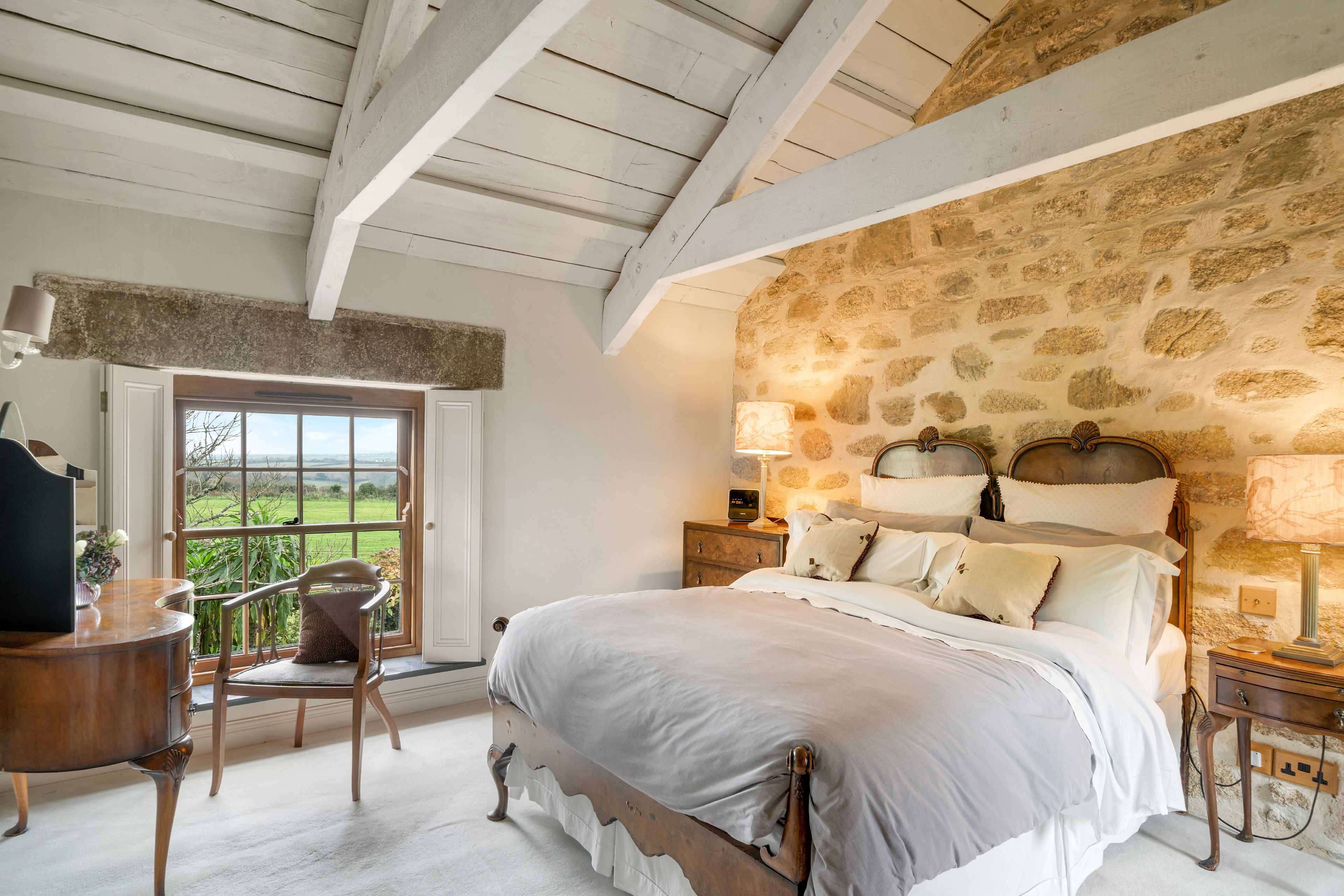 A bedroom with a stone-patterned wall, a wooden bed with white bedding, a nightstand with a lamp, and a chair by a window with a view.