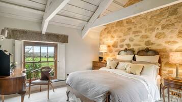 A bedroom with a stone-patterned wall, a wooden bed with white bedding, a nightstand with a lamp, and a chair by a window with a view.