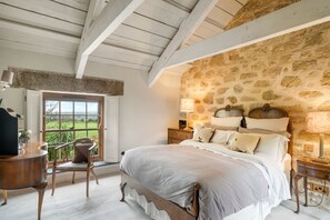 A bedroom with a stone-patterned wall, a wooden bed with white bedding, a nightstand with a lamp, and a chair by a window with a view. - Tregadjack Farmhouse, Tregathenan, Near Porthlevan , Cornwall (Helston)