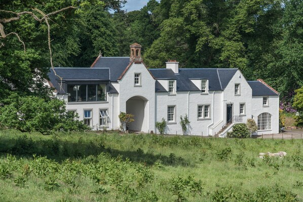 Stately country house with a central archway, surrounded by lush greenery and a well-maintained lawn.