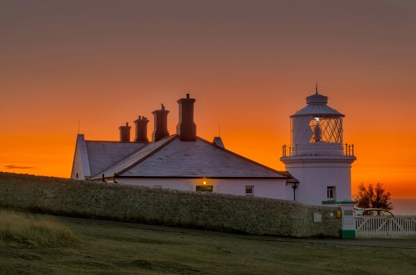 A lighthouse with a white exterior and a lantern room, situated next to a building with multiple chimneys, under a sunset sky.