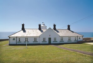 A white building with a gabled roof and multiple chimneys, situated on a grassy area with a clear view of the sea.
