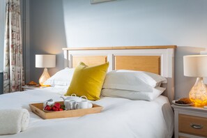 A hotel room with a bed, two bedside lamps, a tray with strawberries and teapots, and a wooden headboard.