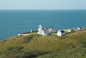 A coastal building with a lighthouse, surrounded by grassy terrain and the sea.