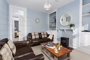 A living room with a brown leather sofa, a wooden coffee table with magazines, a black stove, and a white door leading to another room.