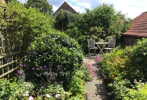 A garden pathway with a wooden fence, various plants, and two white metal chairs.