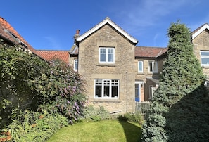 A two-story stone house with a red-tiled roof, a central window, and a small garden area with various plants.