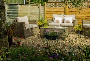 A wicker outdoor seating area with cushions and a table, surrounded by a variety of plants and flowers.