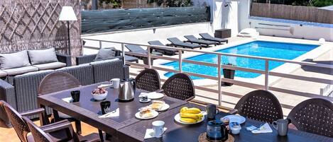 A poolside dining area with a table set for two, a wicker sofa, and a pool in the background.