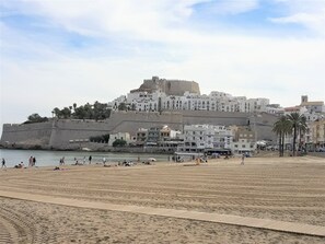 Paisatge urbà costaner amb una imponent estructura semblant a una fortalesa, una platja sorrenca i diversos edificis, incloent un hotel amb múltiples pisos i balcons.
