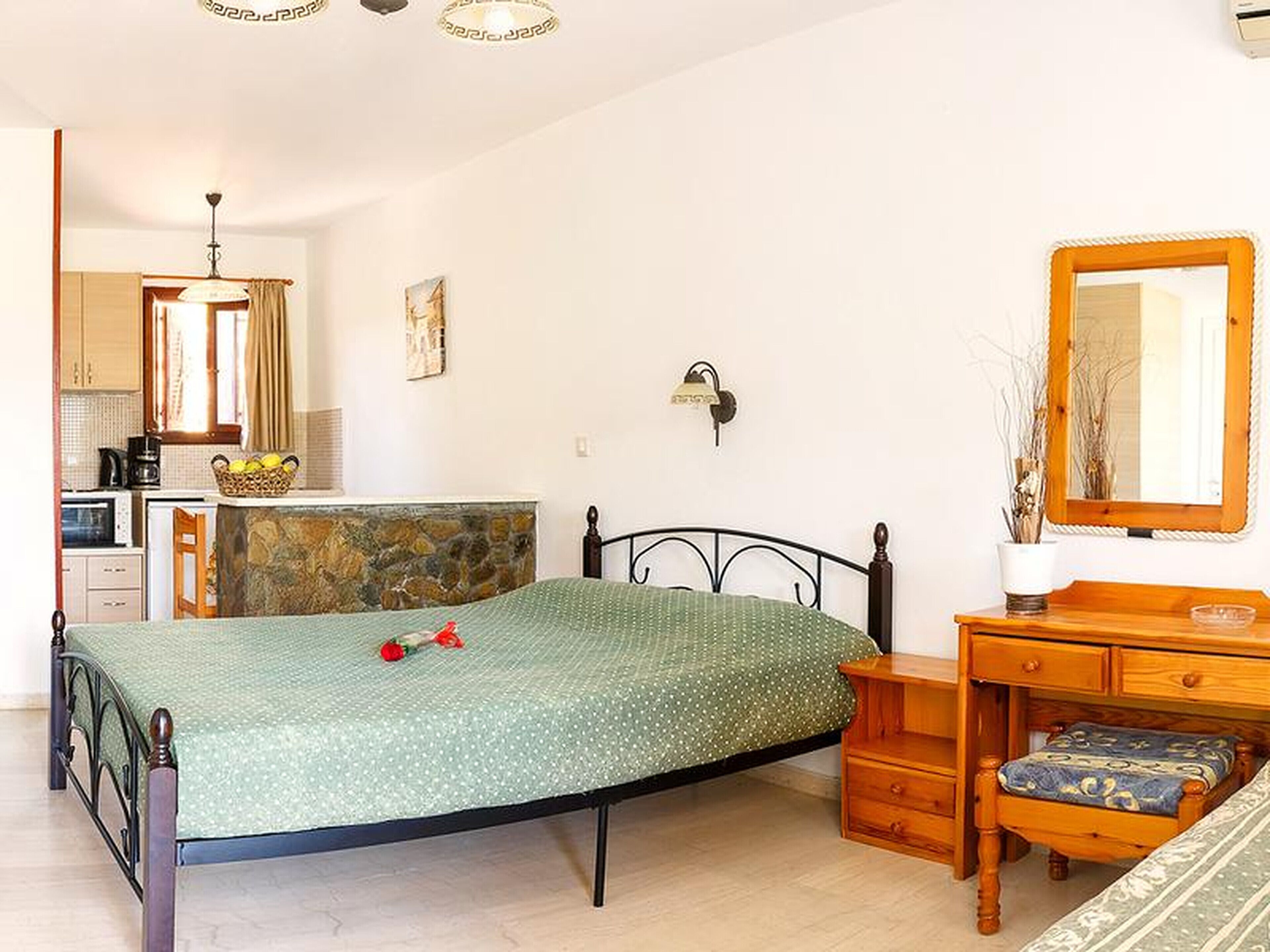 Quarto com cama, cômoda de madeira, espelho e uma cozinha compacta ao fundo.