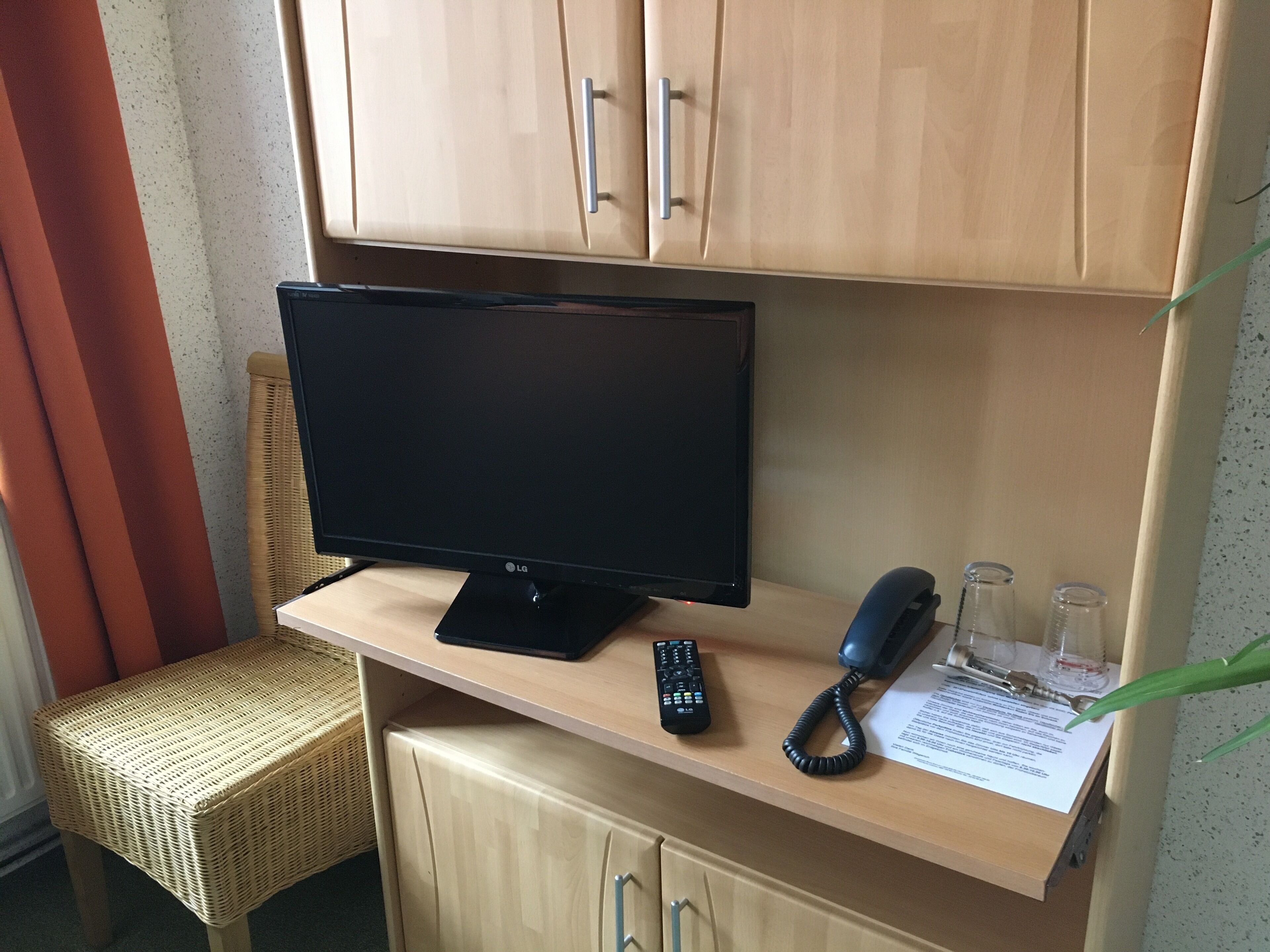 A hotel room with a television on a wooden stand, a remote control, a telephone, and a glass of water on a table.